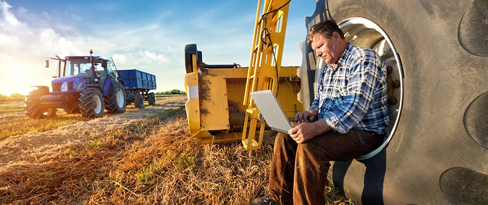 A farmer with agricultural equipment using a laptop computer on his farm